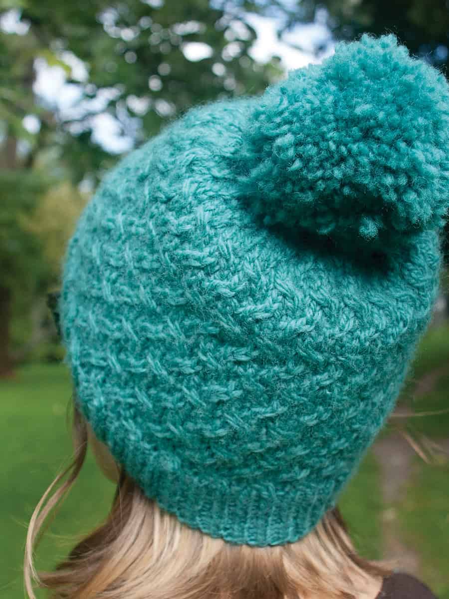 Agnes-cabled-hat-knitting-pattern-3 A person with light brown hair is outdoors, wearing a warm teal hat with a large pom-pom on top. The background shows green grass, trees, and a blurred pathway. The person is facing away from the camera. the design is Agnes worsted weight hat pattern by Faye Perriam-Reed
