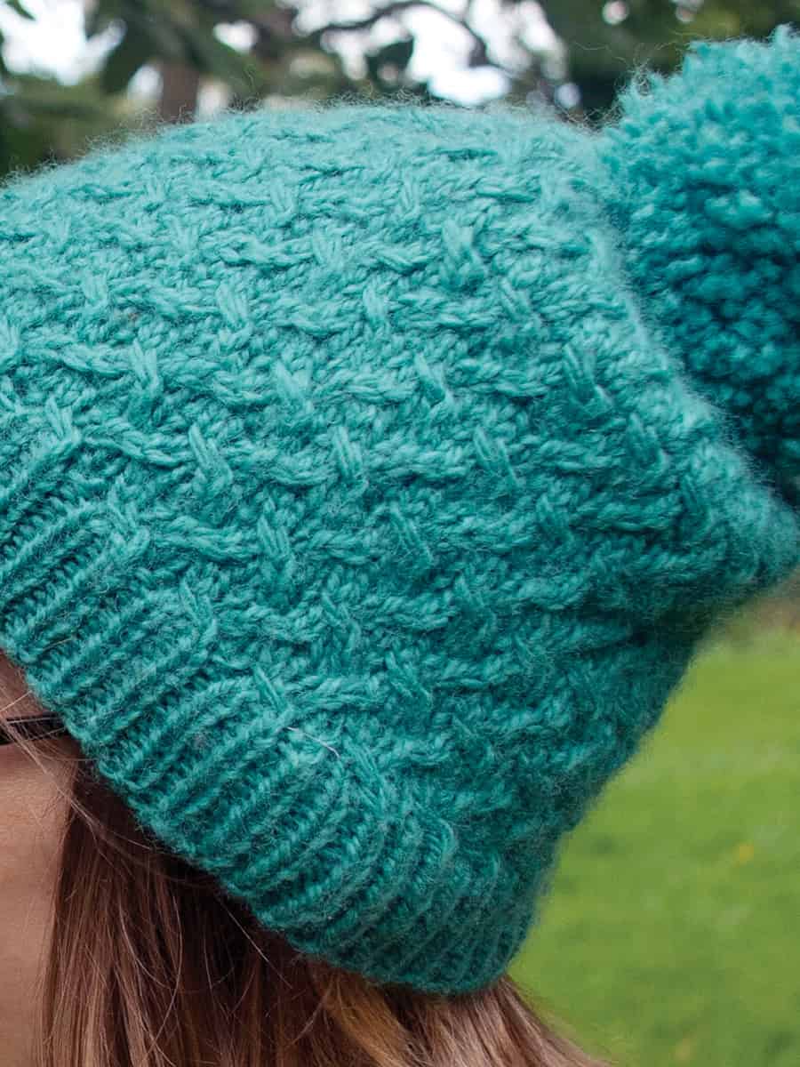Agnes-cabled-hat-knitting-pattern-4 A close-up of a person wearing a knitted teal beanie featuring a cable stitch pattern and a large pom-pom, standing outdoors with green grass in the blurred background.