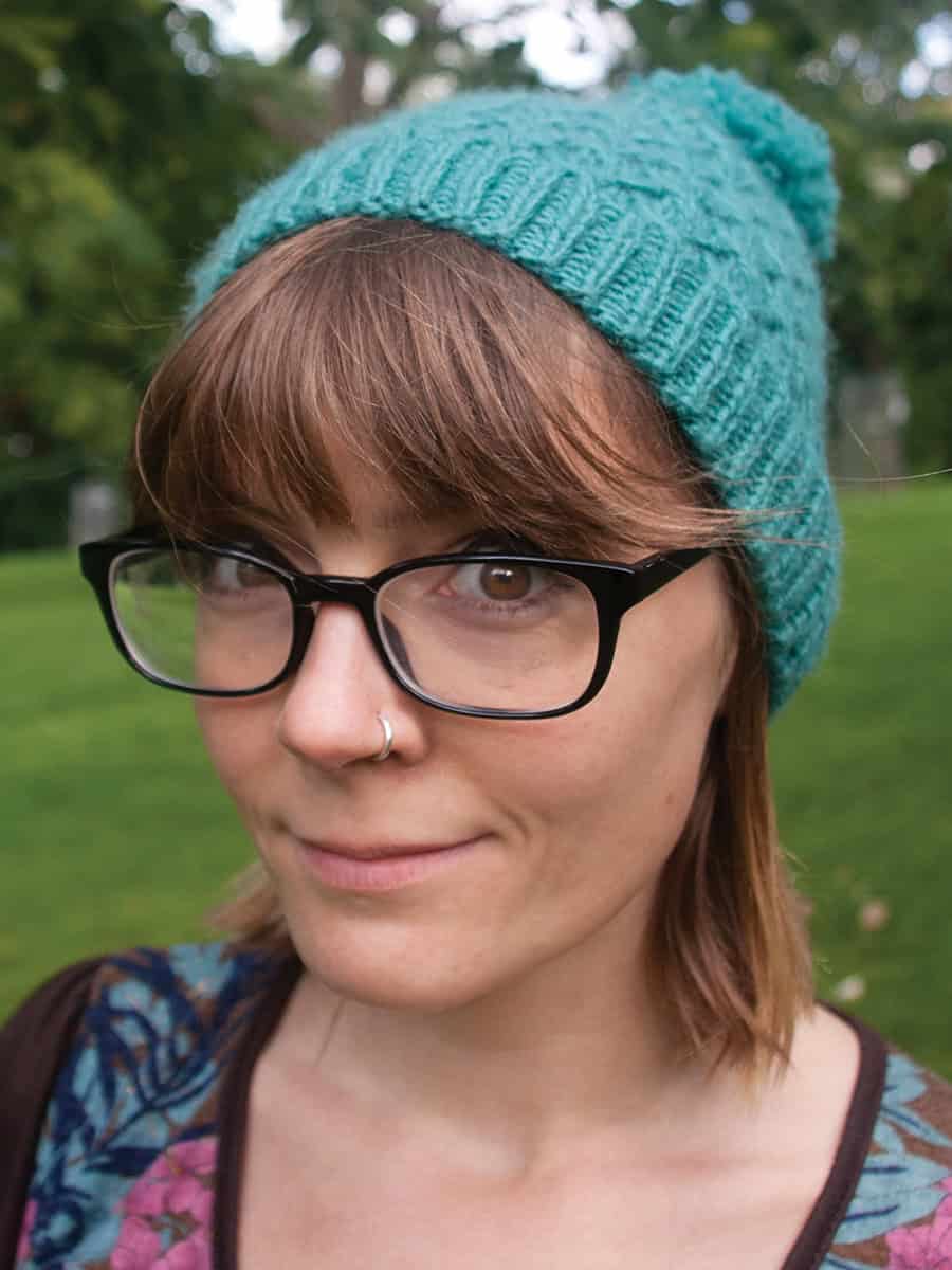 Agnes-cabled-hat-knitting-pattern A person with brown hair and glasses, wearing a turquoise cable stitch beanie and a nose ring, stands outdoors on green grass with trees in the background.