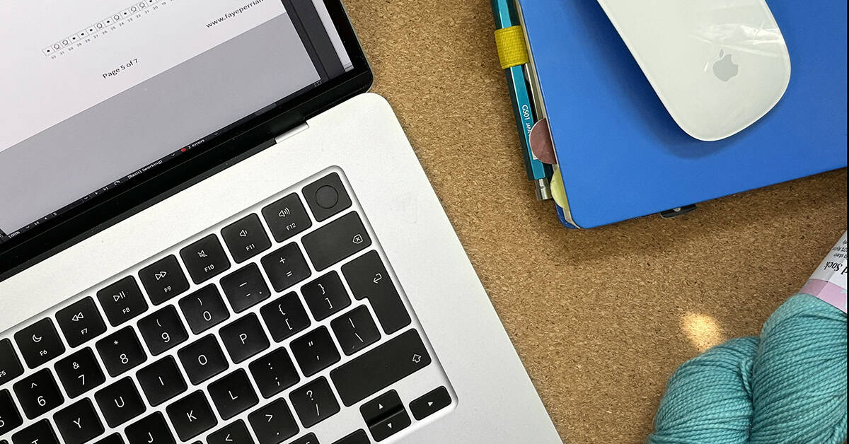 A close-up of a knitting tech editor’s workspace with part of a laptop keyboard, a blue notebook with pen, a white computer mouse, and teal yarn on a cork surface.