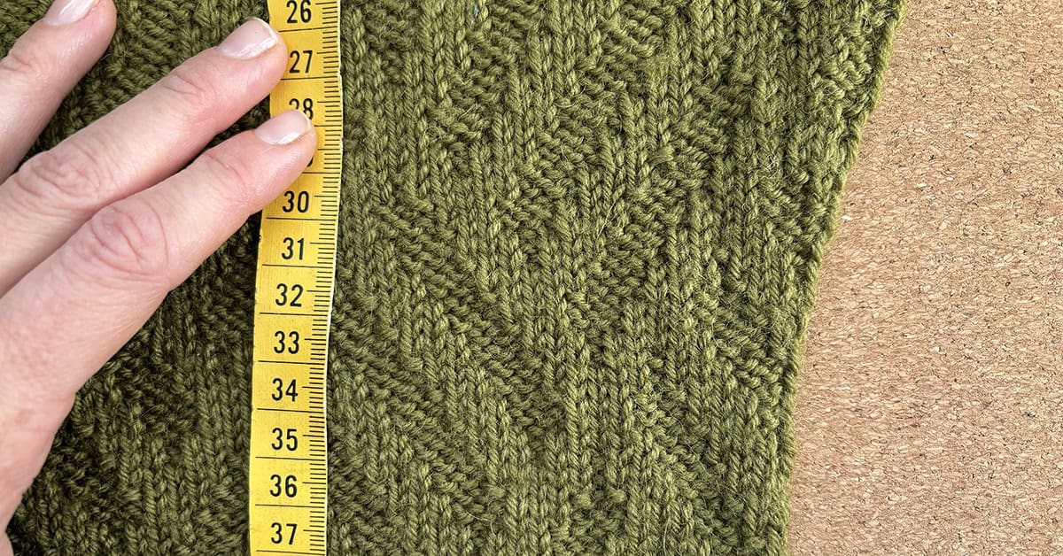 A hand uses a yellow measuring tape to check how long the textured green knitted fabric is—about 31 centimeters—against a cork background, perfect for those looking to tech edit a knitting pattern accurately.