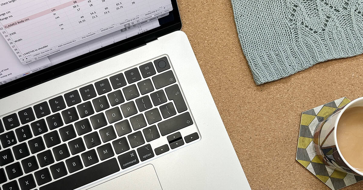 A laptop keyboard and part of the screen showing a spreadsheet sit on a cork surface next to a cup of tea on a patterned coaster and a piece of light blue knitted fabric. Learn how to reduce time with a knitting tech editor in this handy post. 