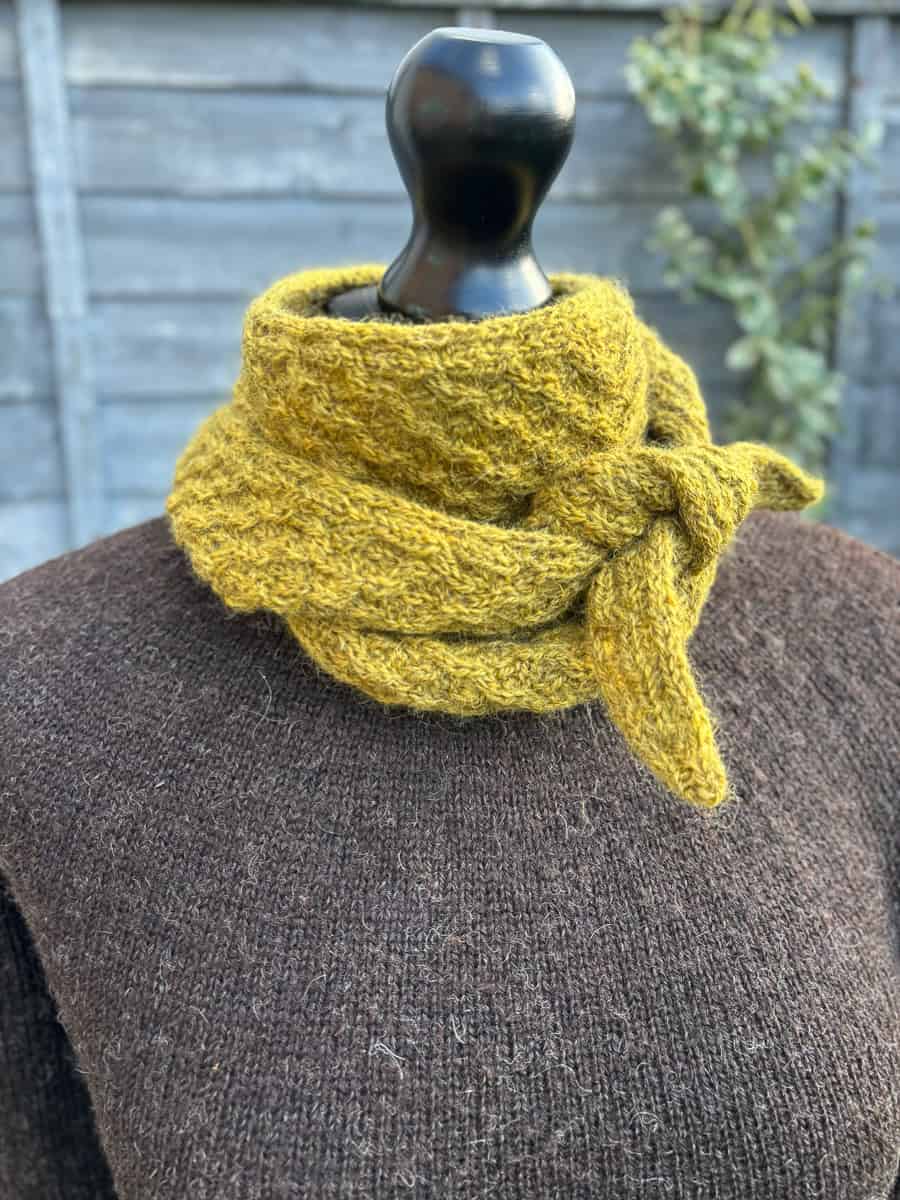 A black mannequin dressed in a brown textured sweater displays a hand-knitted, mustard yellow neck scarf tied in a knot, with a blurred wooden fence and greenery in the background.