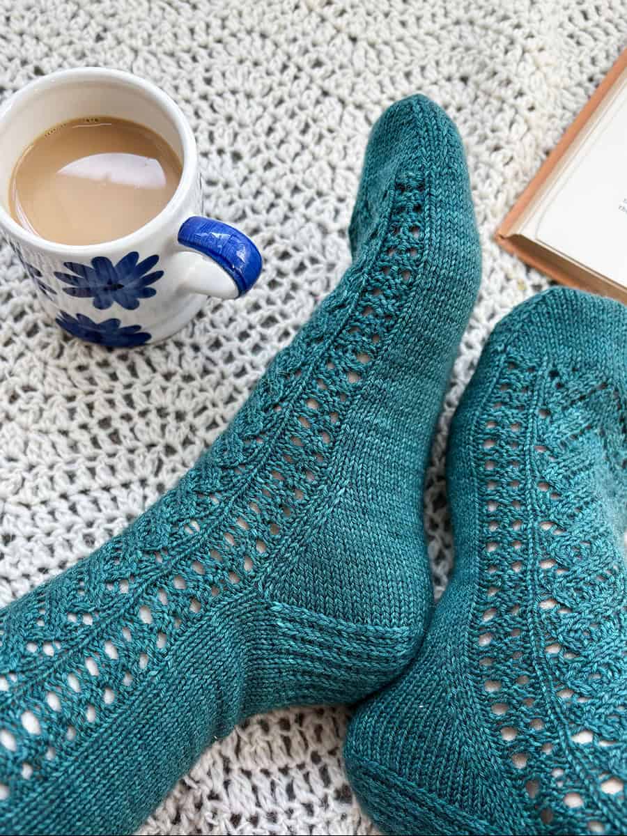 A person wearing teal, lacy knit socks rests on a cream crocheted blanket beside a mug of coffee with blue flower designs and an open book.
