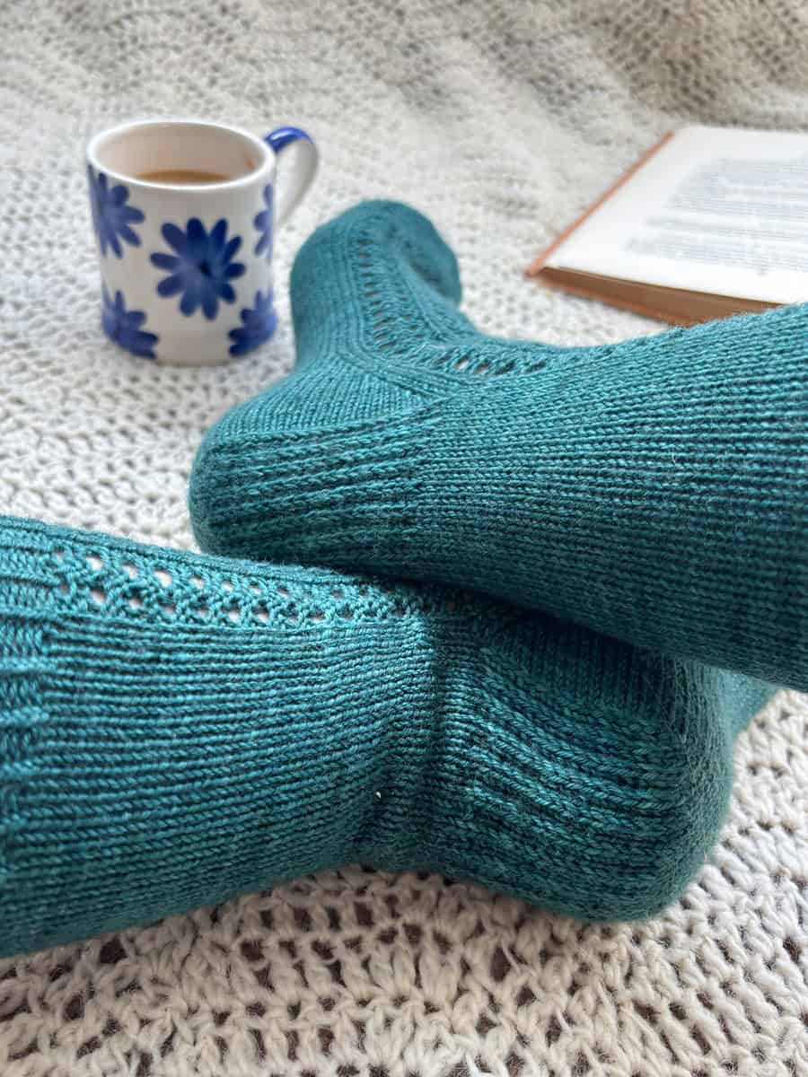 A pair of feet wearing teal knitted socks rest on a cream crocheted blanket. In the background, there is a mug with blue flowers and an open book.