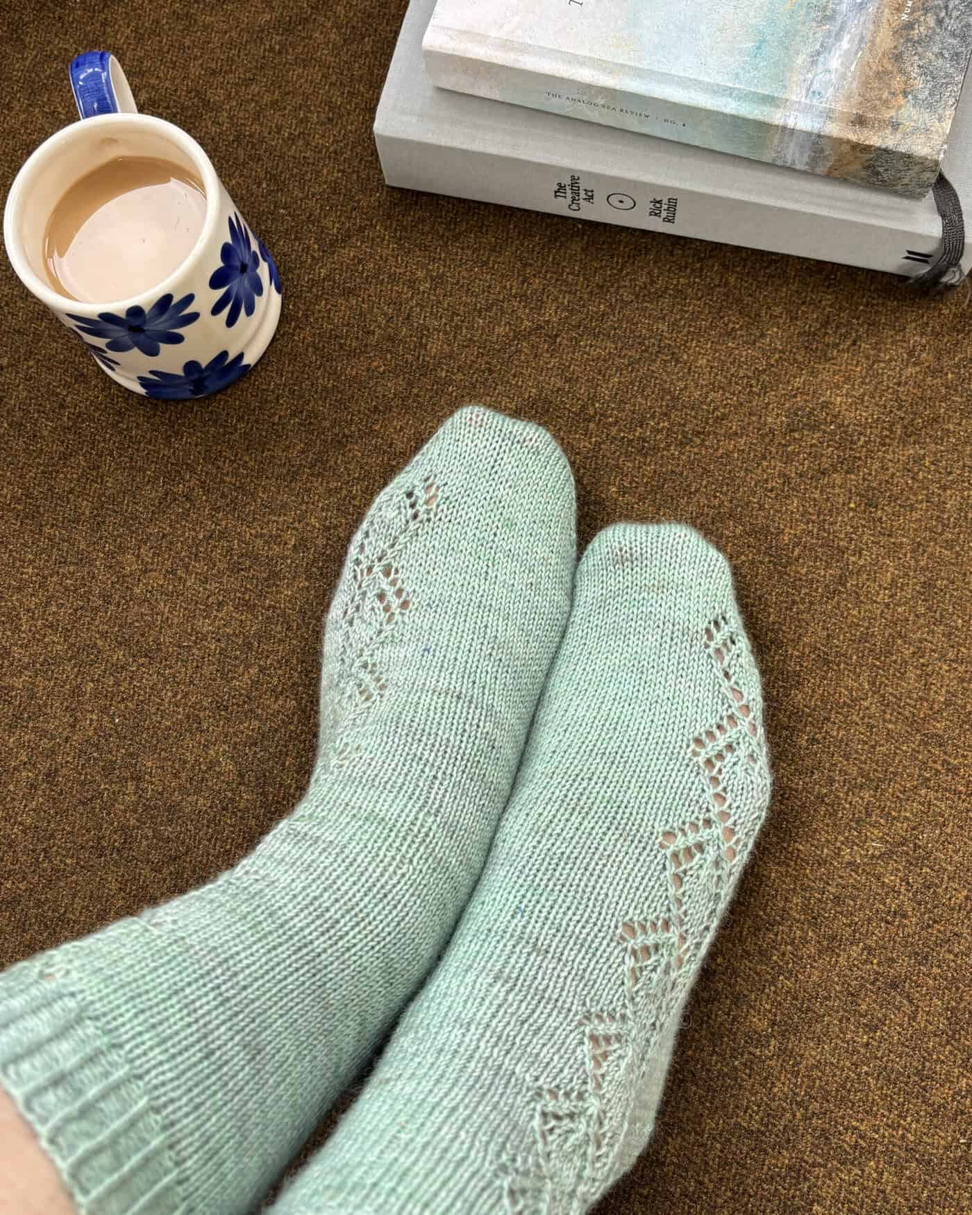 A person wearing mint green cuff down sock pattern knitted socks sits on a brown carpet next to a mug of coffee and a stack of books, creating a cozy and relaxed atmosphere. Pattern is Blisland cuff down sock pattern by Faye Perriam-Reed