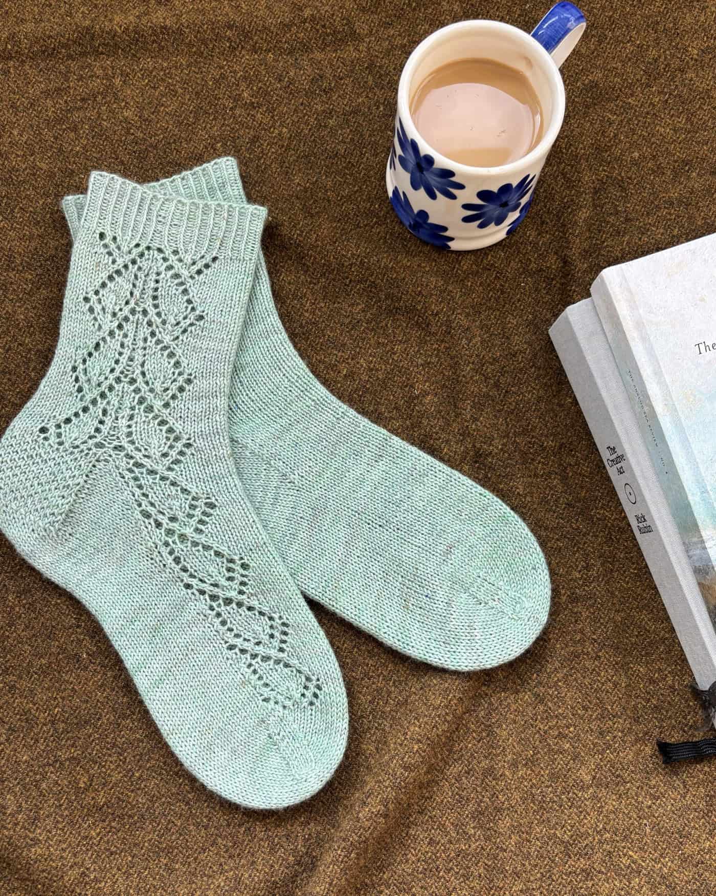 A pair of mint green knitted socks with lace patterns is laid out on a brown blanket beside a cup of tea in a blue flowered mug and two books. The pattern is Blisland cuff down sock pattern by Faye Perriam-Reed