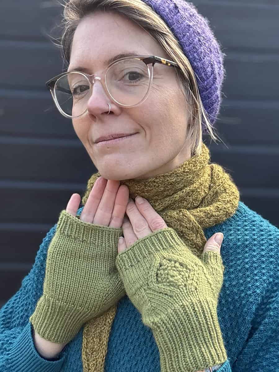 A person wearing glasses, a purple knit hat, a mustard-yellow scarf, a teal textured sweater, and light green fingerless gloves, standing in front of a dark background and smiling gently.
