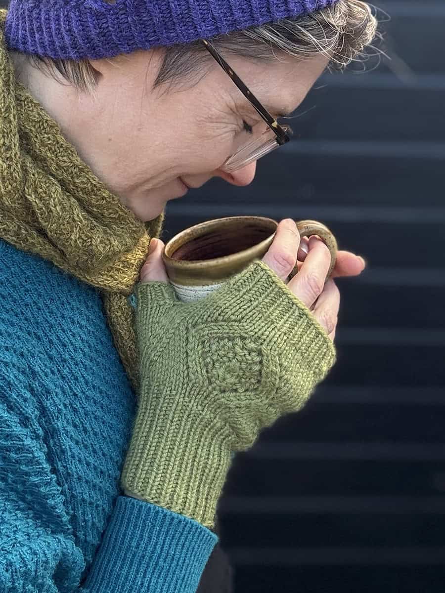 A person wearing a purple hat, green scarf, teal sweater, and green fingerless gloves holds a ceramic mug close to their face, appearing to enjoy its warmth. The background is dark and blurred.