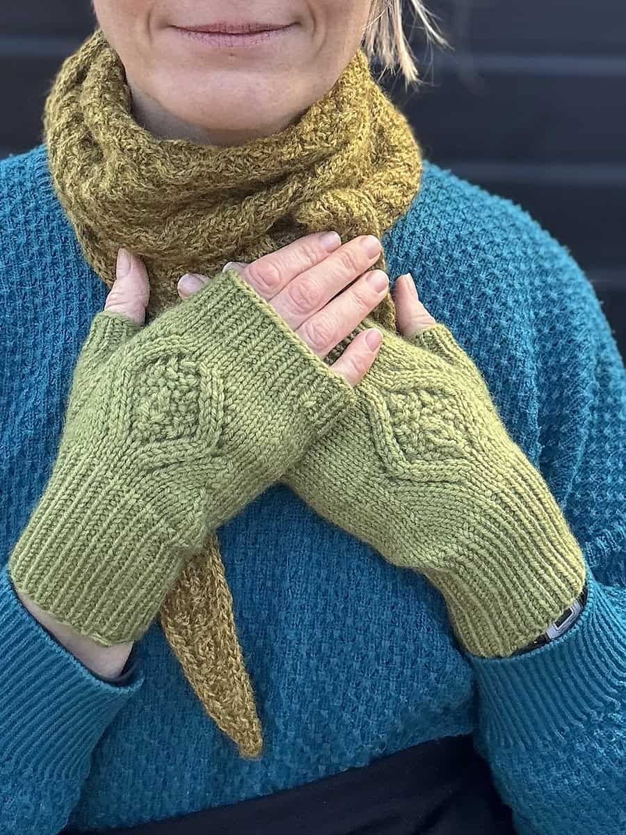 A person wearing a textured teal sweater, a knitted mustard yellow scarf, and light green mitts with a diamond pattern motif holds their hands over their chest. Pattern is Merry Maidens DK easy mitts pattern by Faye Perriam-Reed