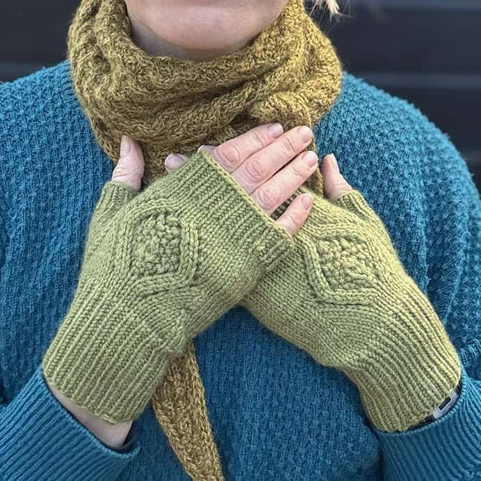 A person wearing a teal sweater and a textured mustard scarf displays their hands, which are covered by light green knitted mitts with a diamond motif on the back of the hand. 