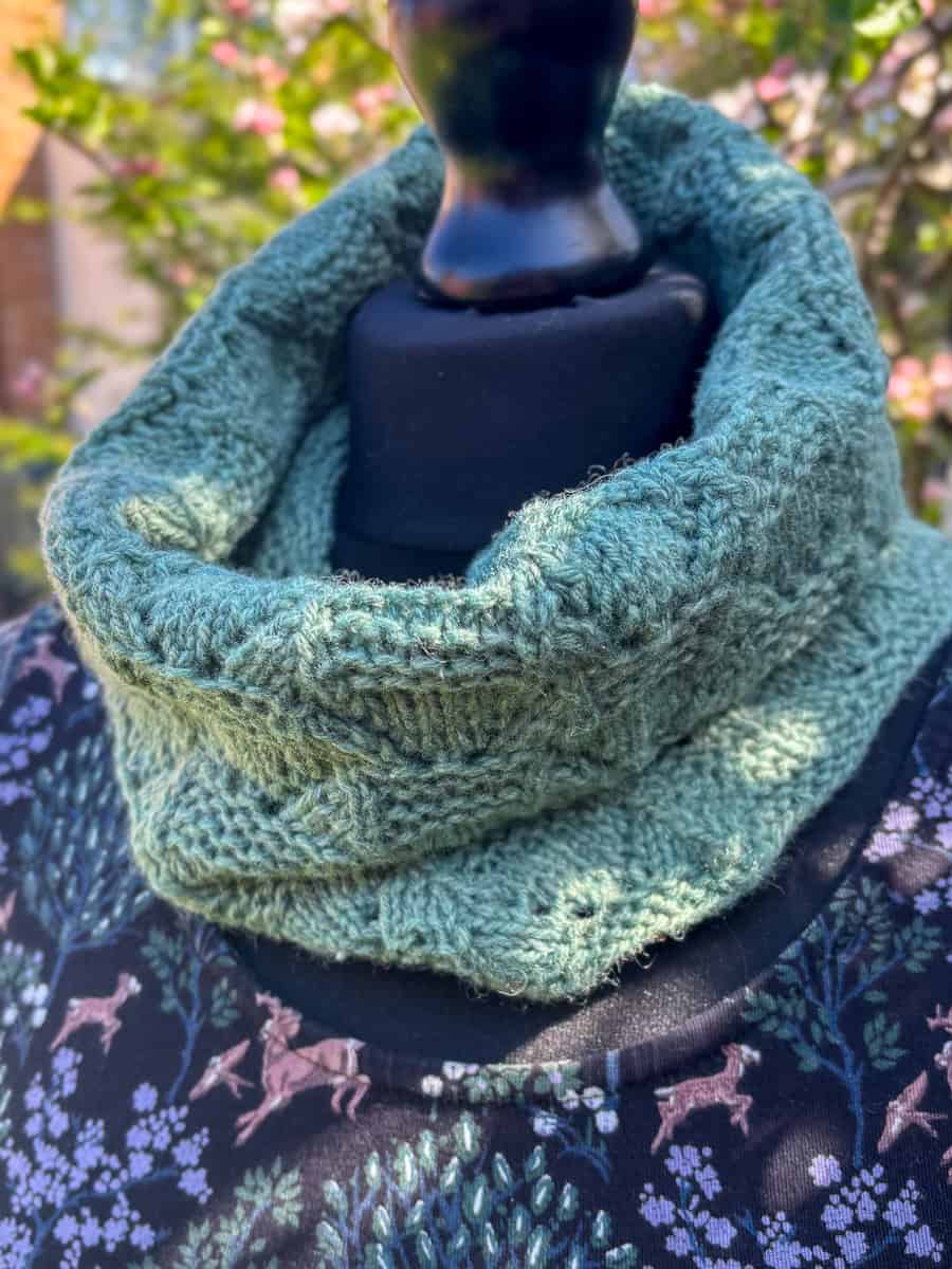 A textured, light green knitted cowl is displayed on a black mannequin outdoors, with blurred greenery and pink blossoms in the background. The mannequin wears a patterned top with trees and deer.