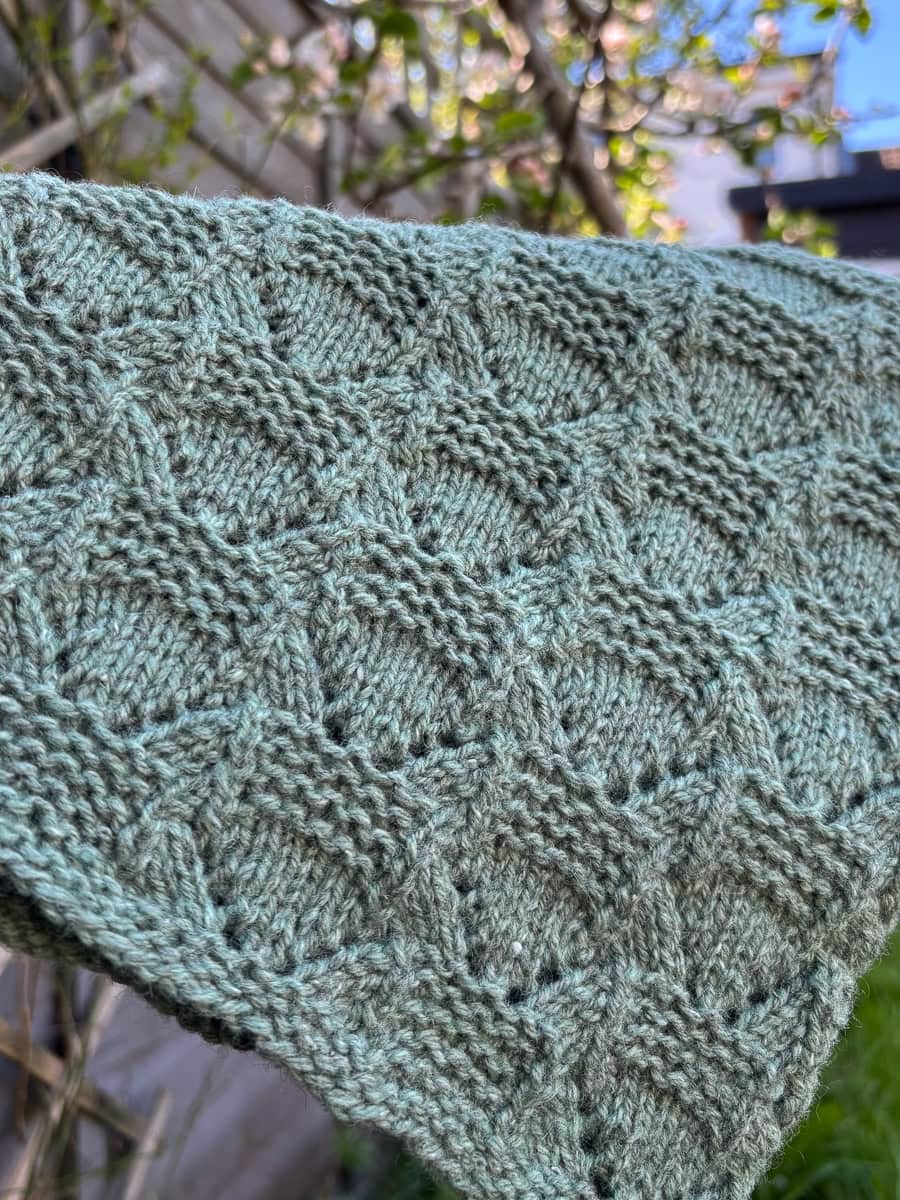 A close-up of a green knitted cowl with a geometric lace and garter stitch pattern, held outdoors with blurred greenery