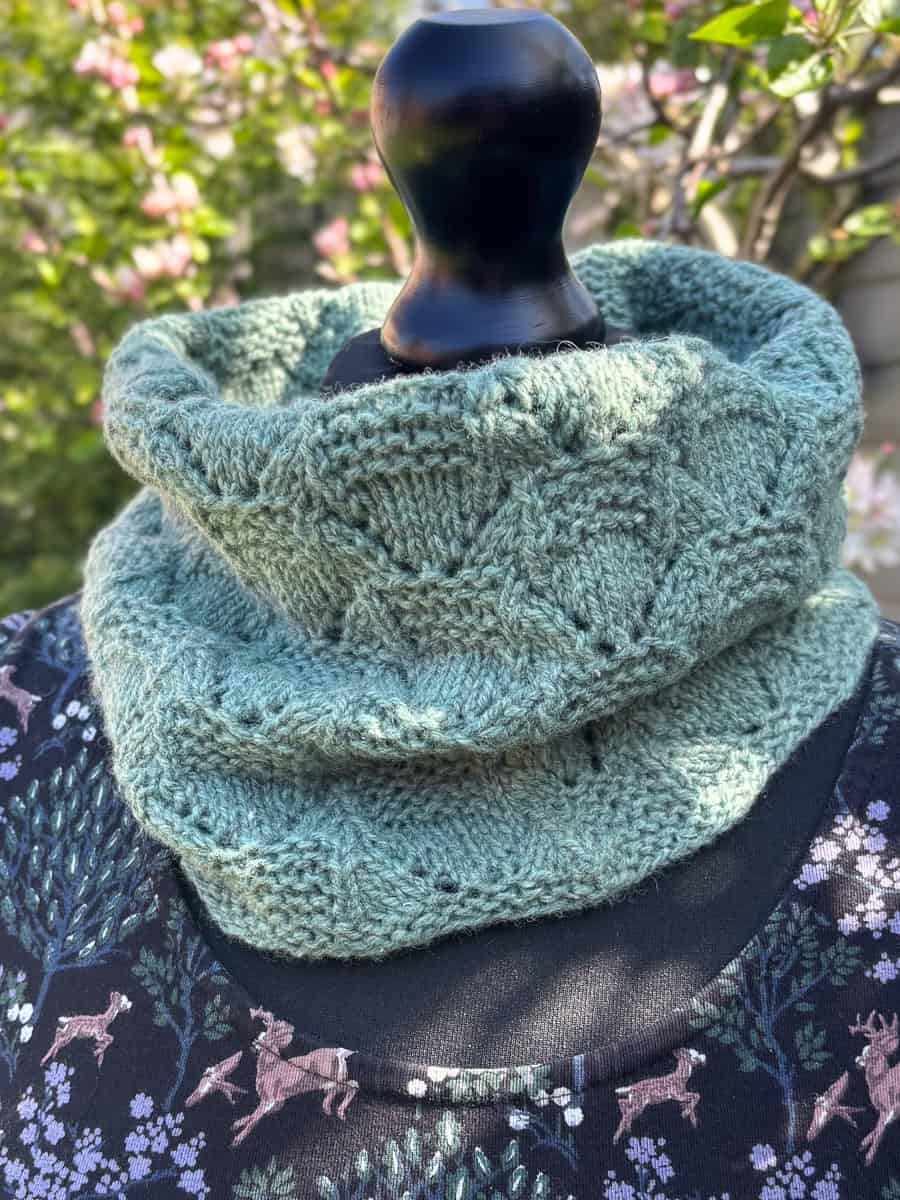 A knitted light green cowl with a lace and garter stitch pattern is displayed on a dressmaker’s mannequin outdoors, with a floral and deer patterned dress and blooming branches in the background.