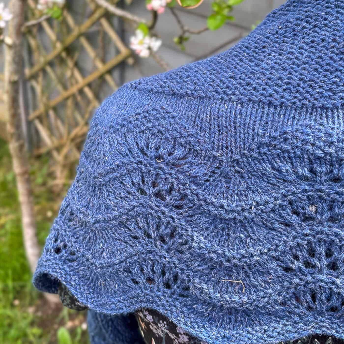 Close-up of a blue knitted shawl, featuring a feather and fan lace pattern and garter stitch, draped over a mannequin's shoulders. The background features blurred greenery, a wooden trellis, and blooming flowers.