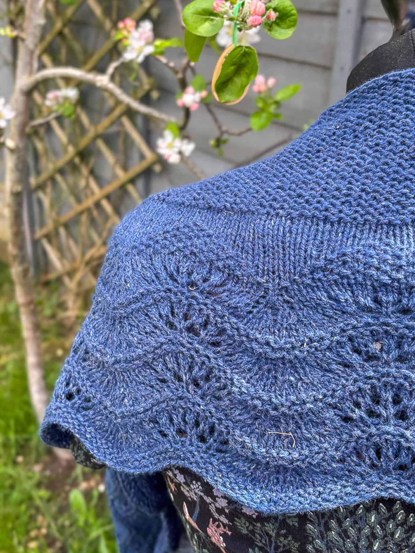 A close-up of a blue knitted shawl in a simple garter stitch and feather and fan pattern, shown draped over a mannequin outdoors near blossoming branches and a wooden fence.