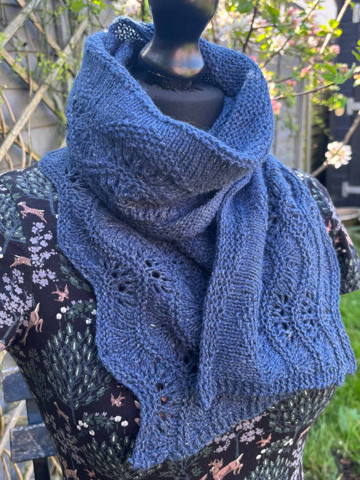 A mannequin wears a textured, hand-knitted blue shawl with wavy edges and a feather and fan lace pattern, styled around the neck. The mannequin is outdoors, dressed in a floral-patterned top, with greenery and trellis in the background.