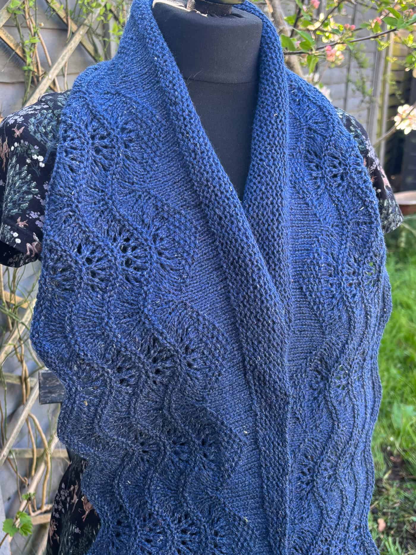 A dark blue knitted scarf featuring garter stitch and a lace feather and fan pattern, displayed on a mannequin outdoors with a floral dress and greenery in the background.