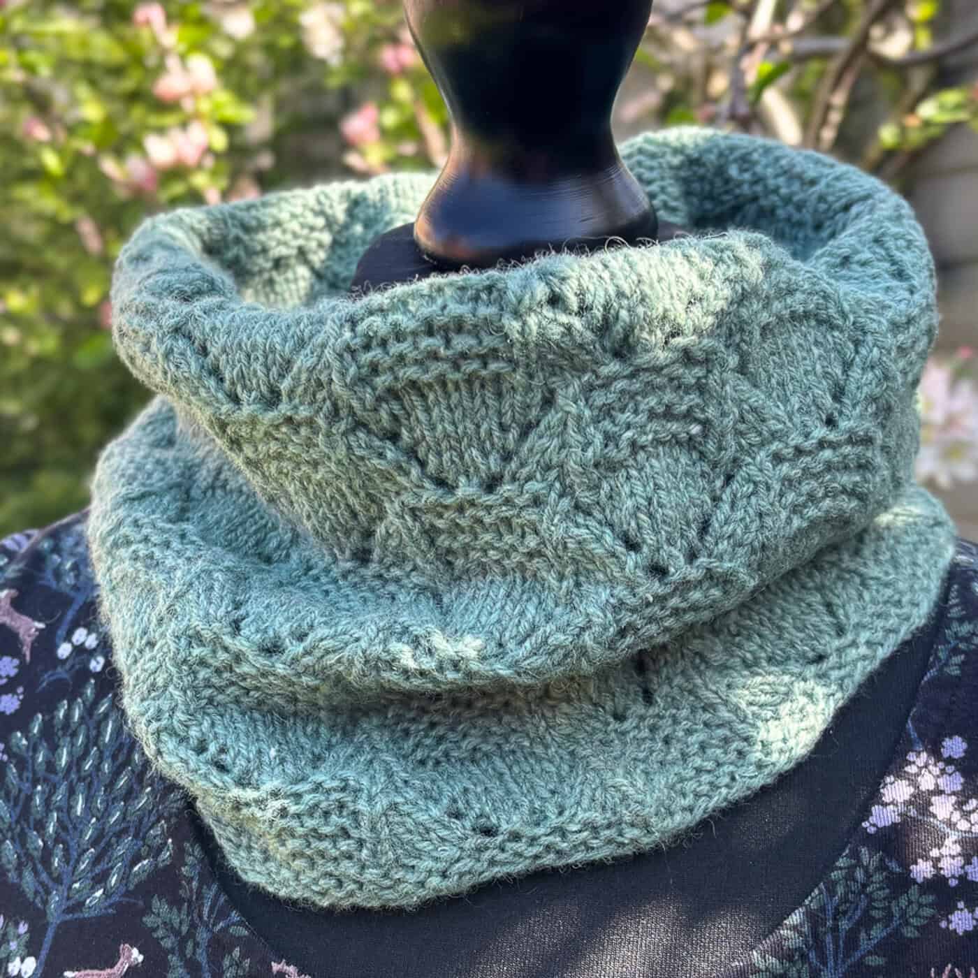 A light green hand-knitted cowl, perfect for one skein patterns, features a textured geometric design and is displayed on a black mannequin outdoors amid blurred greenery and pink flowers in the background.