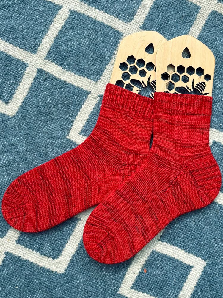 Two bright red knitted socks are stretched on wooden sock blockers with honeycomb cut-outs, placed side by side on a blue and white patterned rug.