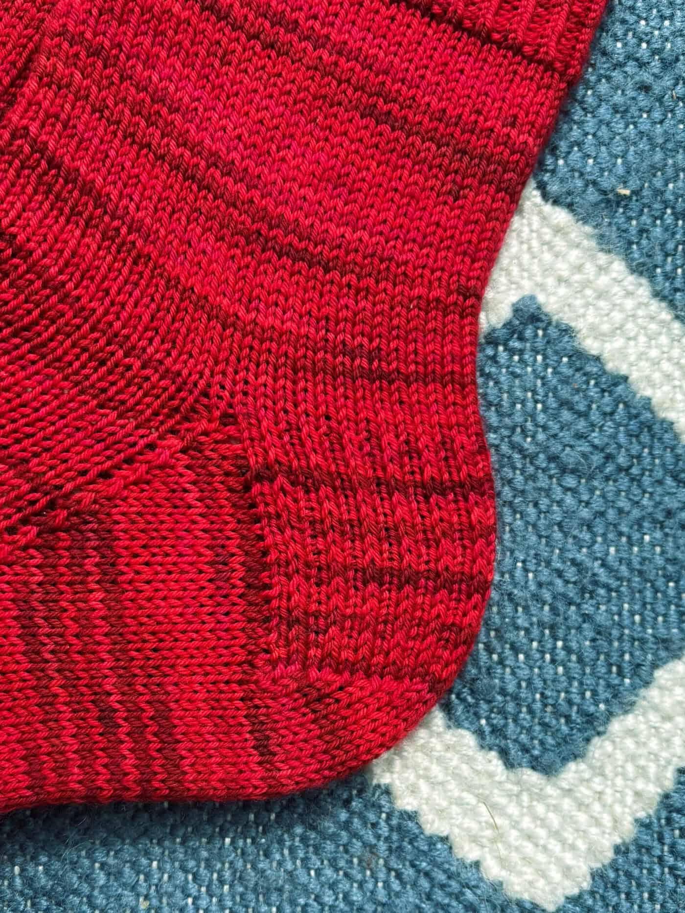A close-up of a red and dark red striped knitted sock on a blue and white patterned textured rug.