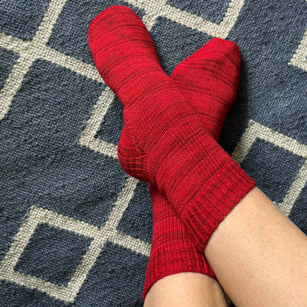 Feet wearing bright red socks—crafted from one skein patterns—are crossed at the ankles, resting on a blue and white geometric patterned rug.