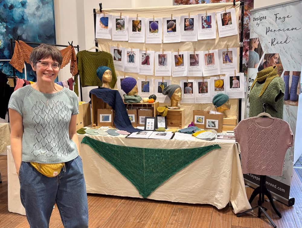 A woman standing at a craft stall displaying hand-knitted jumpers, shawls, and printed pattern sheets for sale.