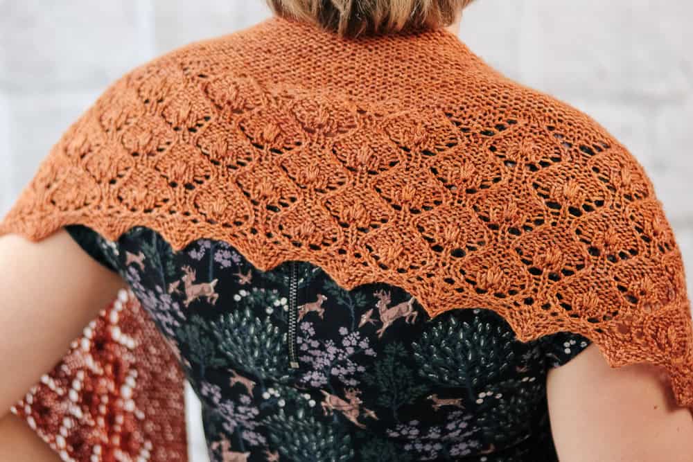 A rust-coloured, hand-knitted shawl with an intricate lace pattern draped over the shoulders of a person wearing a forest-themed print dress.