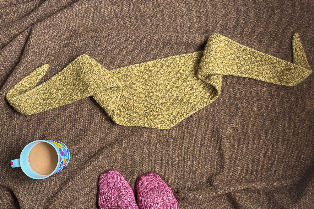 A green knitted triangular shawl, a mug of coffee, and a pair of pink knitted socks resting on a brown blanket.