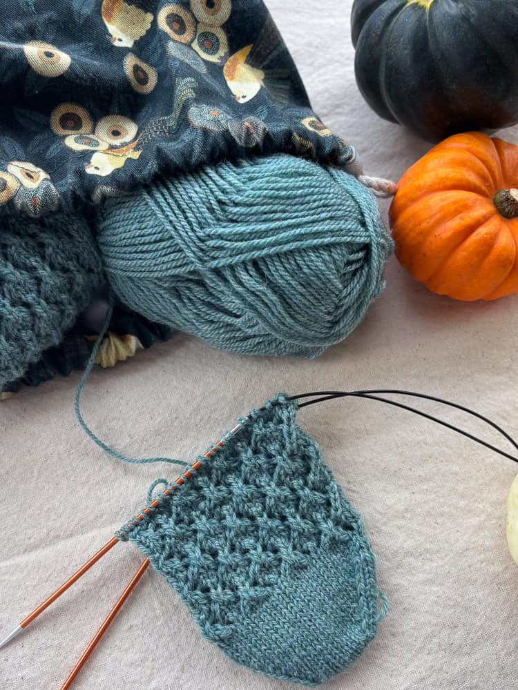 A ball of teal wool and a partially knitted project on circular needles, next to two small pumpkins.